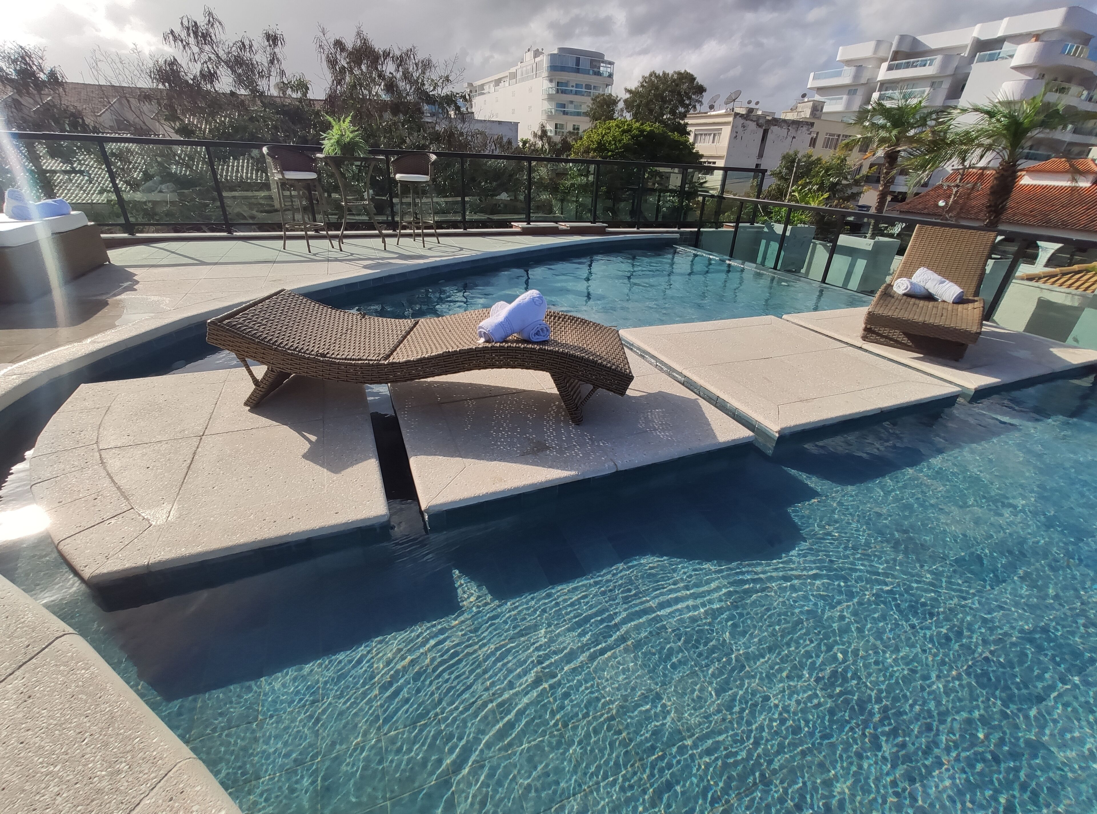 A swimming pool with lounge chairs and a small table, surrounded by a glass railing.