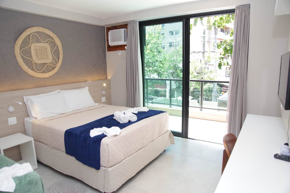 Une chambre d’hôtel équipée d’un lit, d’un bureau, d’une chaise, d’une télévision et d’un balcon offrant une vue sur les bâtiments et les arbres.