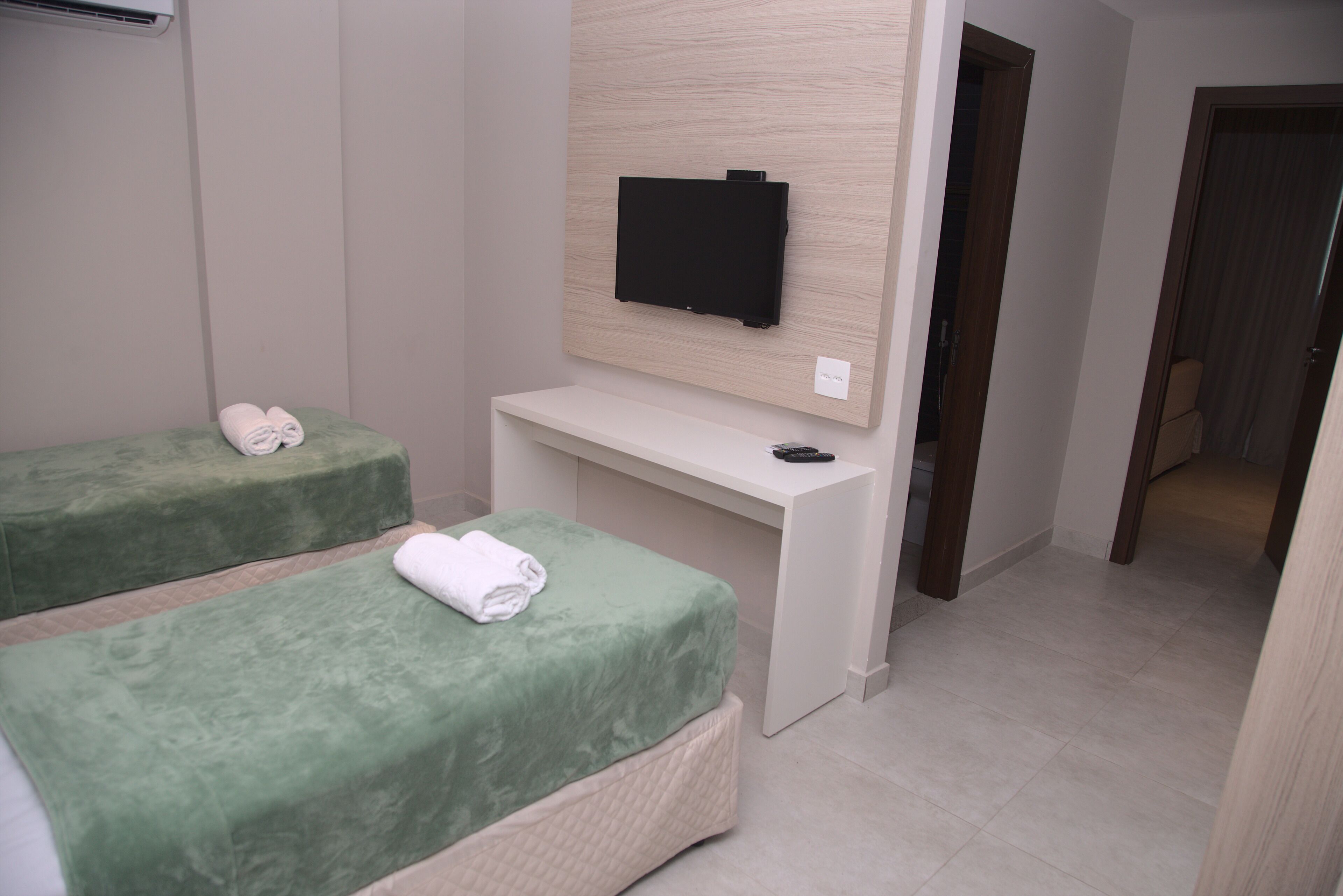 Quarto com duas camas verdes, uma escrivaninha branca, televisão de tela plana na parede e uma porta que dá para outro cômodo.
