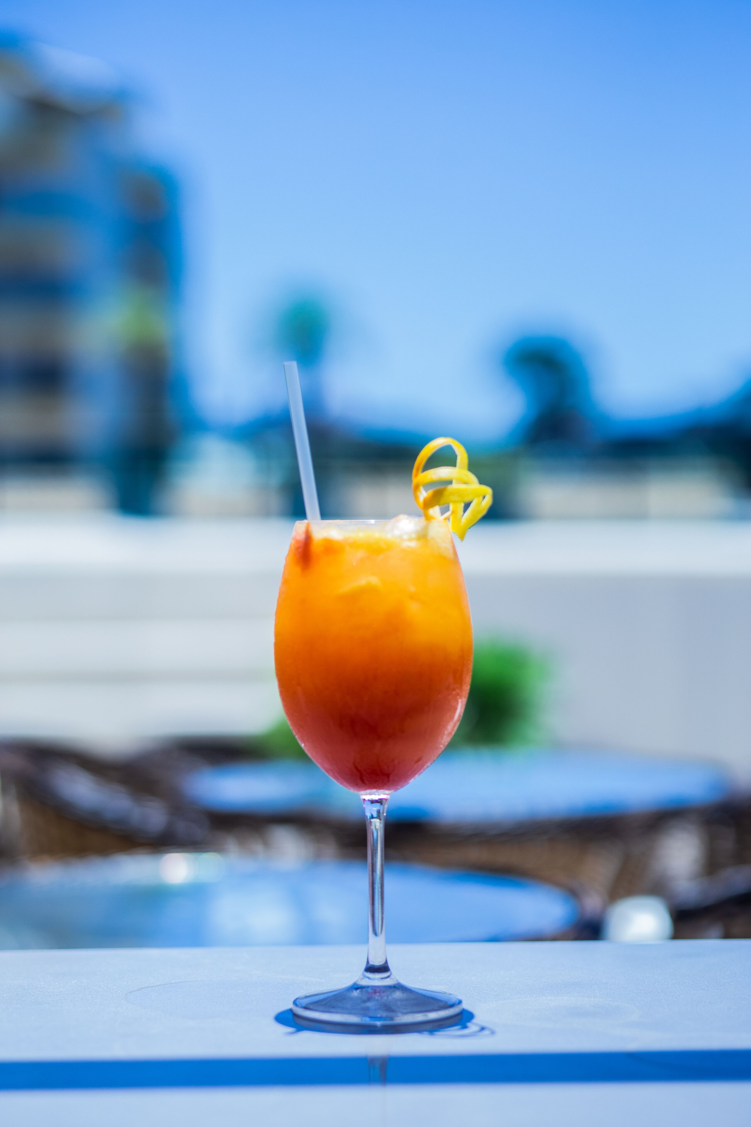 A cocktail in a stemmed glass with a straw and a lemon twist garnish, placed on a table with a blue surface.