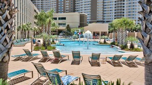 Zona de piscina de un resort con sillones de descanso, una fuente central y edificios altos al fondo.