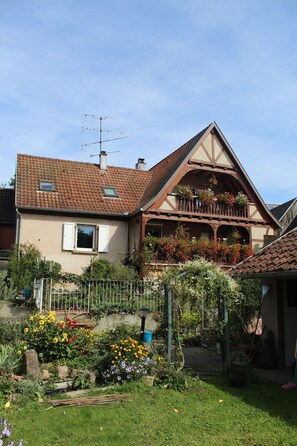 Et traditionelt hus med skråtag, en balkon med blomster og en have med forskellige planter og blomster.
