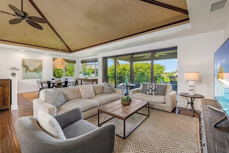 A spacious living area with a large sofa, a coffee table, and a TV. There is a ceiling fan, a dining area with a table and chairs, and large windows offering a view of greenery outside.