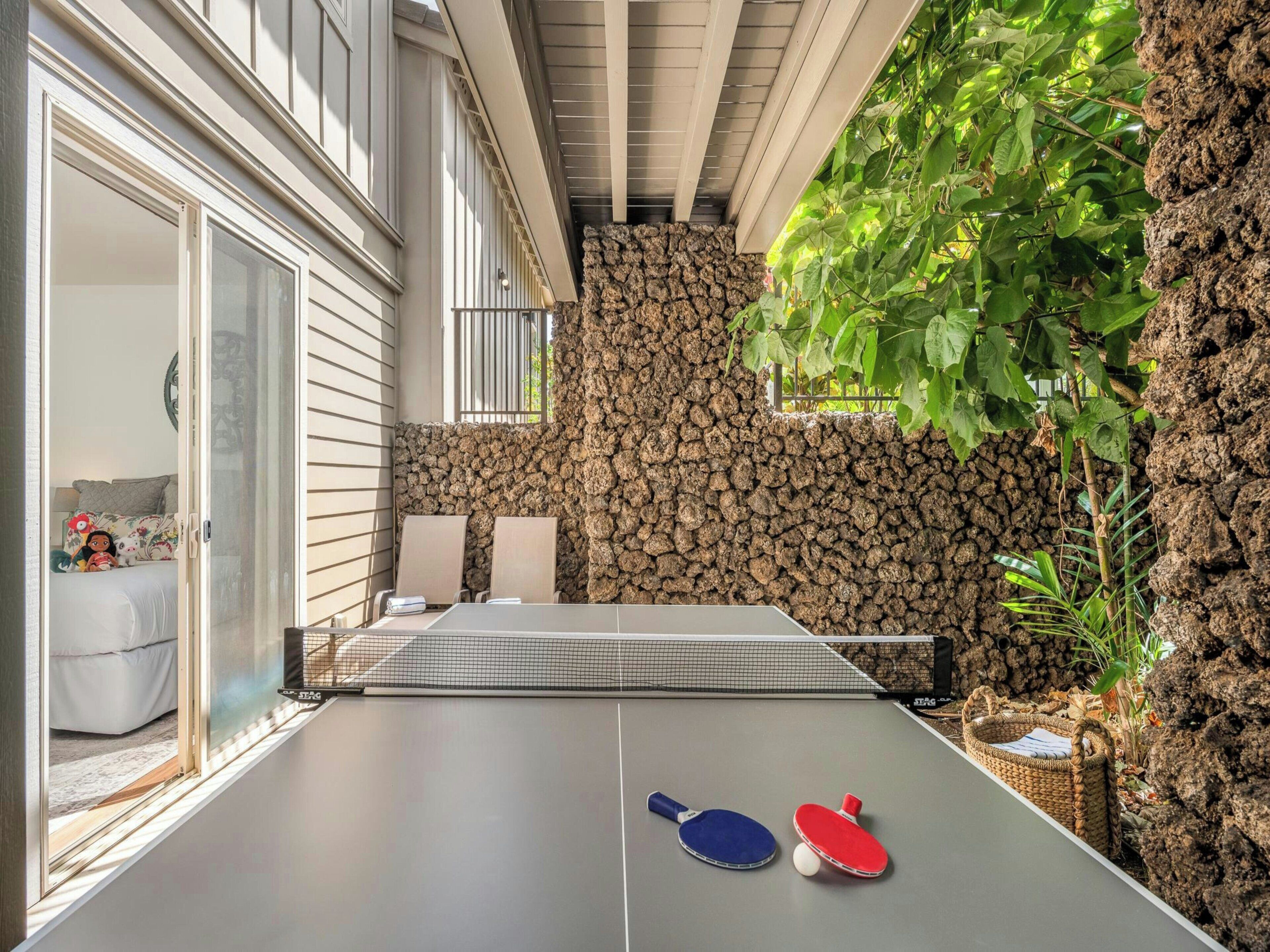 Une table de ping-pong d’intérieur, avec deux raquettes et une balle, un mur en pierre et une vue sur une pièce avec un lit et une fenêtre.