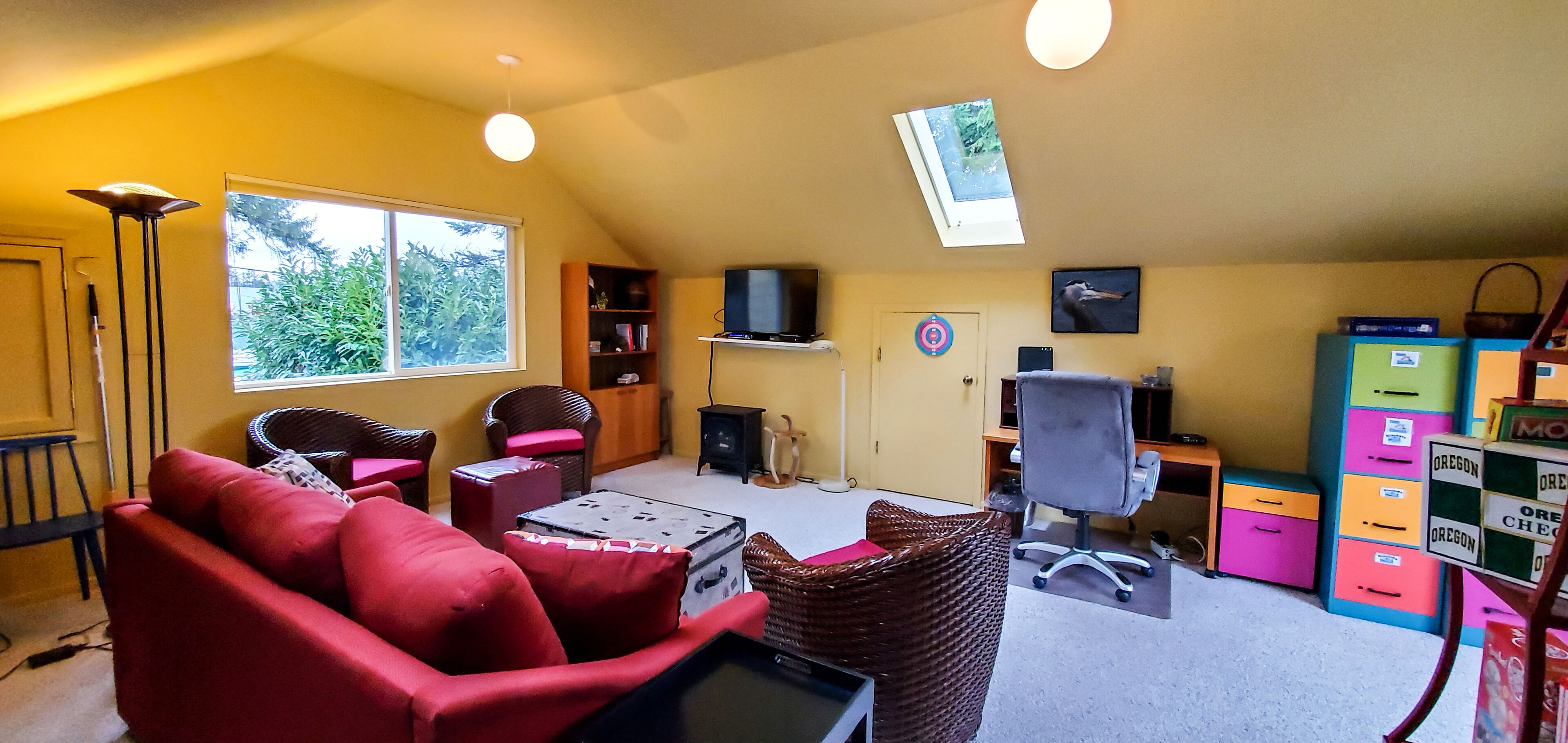 Une pièce chaleureuse avec un canapé rouge, un fauteuil en osier, un bureau, un ordinateur, une télévision, une bibliothèque et une fenêtre donnant sur un paysage verdoyant.