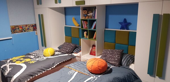A bedroom with two beds, each with a distinct patterned bedspread. There are pillows with various designs, a shelf with books and toys, and a wall with a colorful mural.