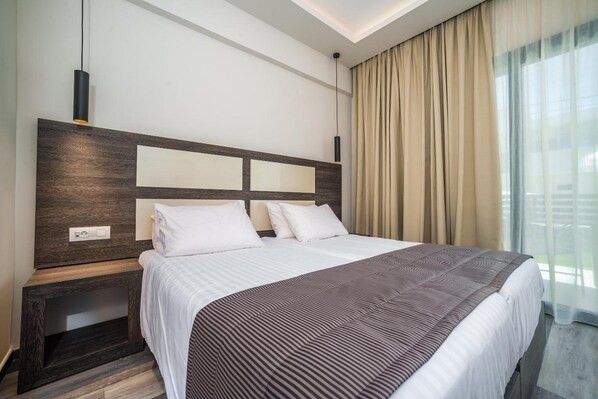 A modern hotel room with a large bed, a wooden headboard, a bedside table, and a window with curtains. - Abaton Luxury Resort (Zakynthos)