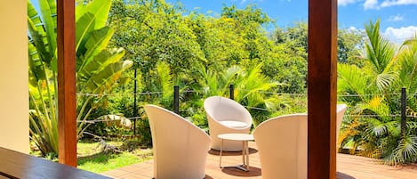 Un área de estar al aire libre con terraza de madera, dos sillas blancas y abundante vegetación de fondo.