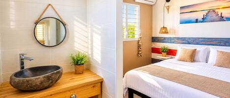 A bathroom with a stone sink, wooden vanity, and a mirror. The room has a bed with white bedding and a painting of a sunset over water.