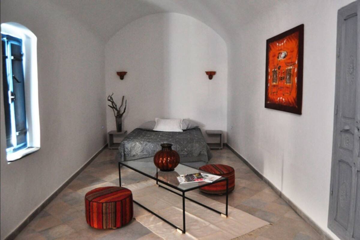 Un couloir étroit mène à une chambre où se trouvent un lit, un vase et une table avec un magazine.