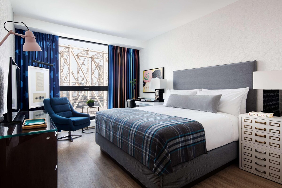 A hotel room with a large bed, a desk with a lamp, a chair, and a view of a bridge.