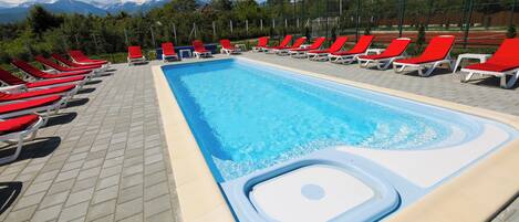 A swimming pool with red and white lounge chairs, a hot tub, and a mountainous backdrop.