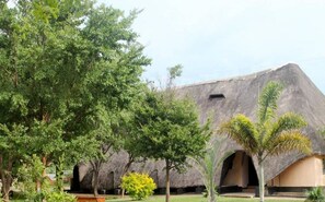 A building with a unique thatched roof and arched openings, surrounded by lush greenery and trees. - Shamkan Lodge (Lusaka)