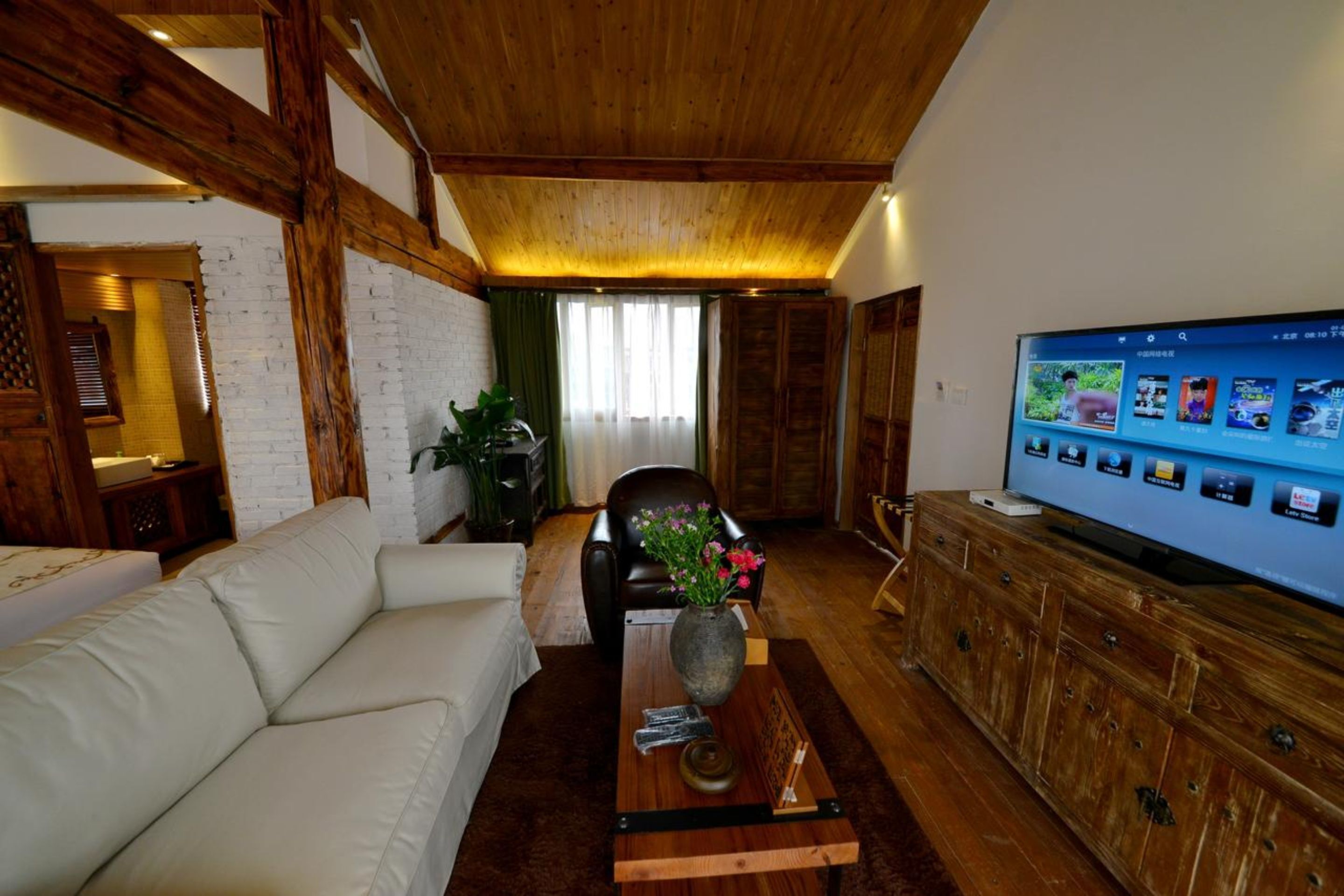 Un accogliente soggiorno con soffitto in legno, un divano bianco, una poltrona in pelle, un mobile in legno e una televisione che mostra un menù.