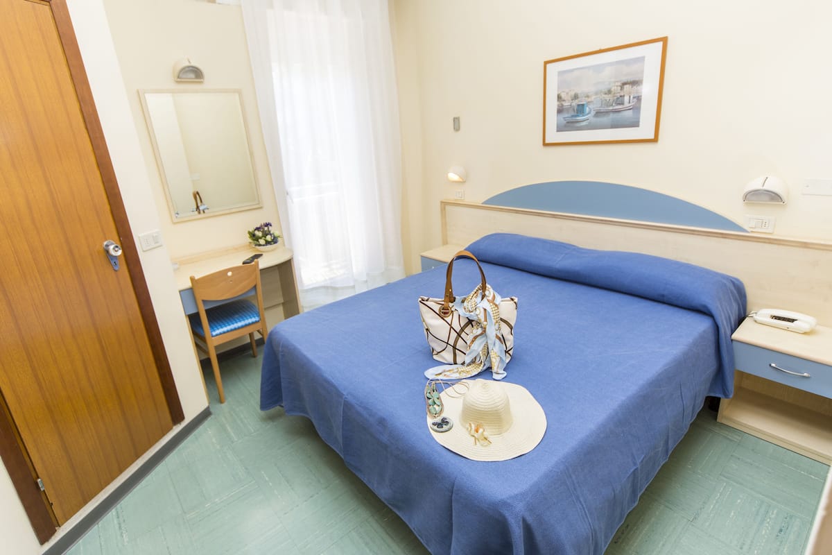 A hotel room with a bed, a bedside table, a chair, a desk, a mirror, and a framed picture on the wall.