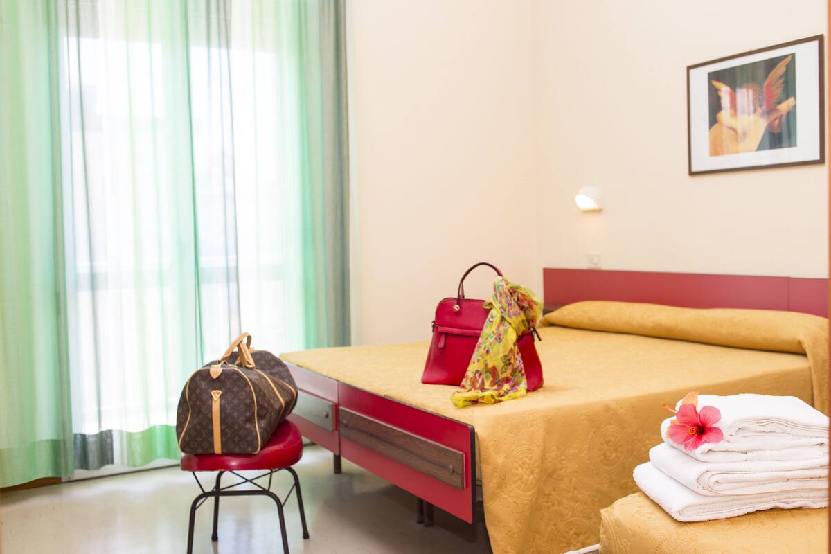 A hotel room with a bed, a red chair, a suitcase, a handbag, and a folded towel with a flower.