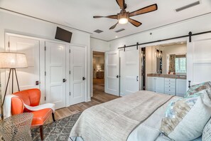 Quarto com cama, cadeira, ventoinha no teto e porta deslizante para um banheiro.