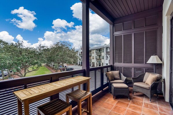 Uma varanda com mesa e cadeiras de madeira, vista para uma rua arborizada e um edifício com persianas.