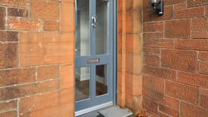 A brick building with a grey door, a small step leading up to the entrance, and a wall-mounted light fixture. - Troon Golf 2024 (Troon)