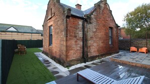 A brick building with a sloped roof, a patio with a wooden table and chairs, and a grassy area with a fence. - Troon Golf 2024 (Troon)