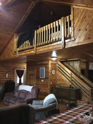 A rustic cabin interior with a loft bedroom, wooden walls, a checkered tablecloth, and a staircase.