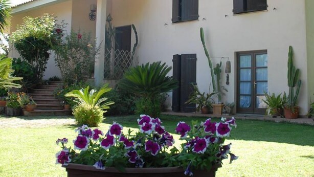 A house with a covered porch, a front door, and a garden with potted plants and flowers. - B&B Fra e Fe (Arborea)