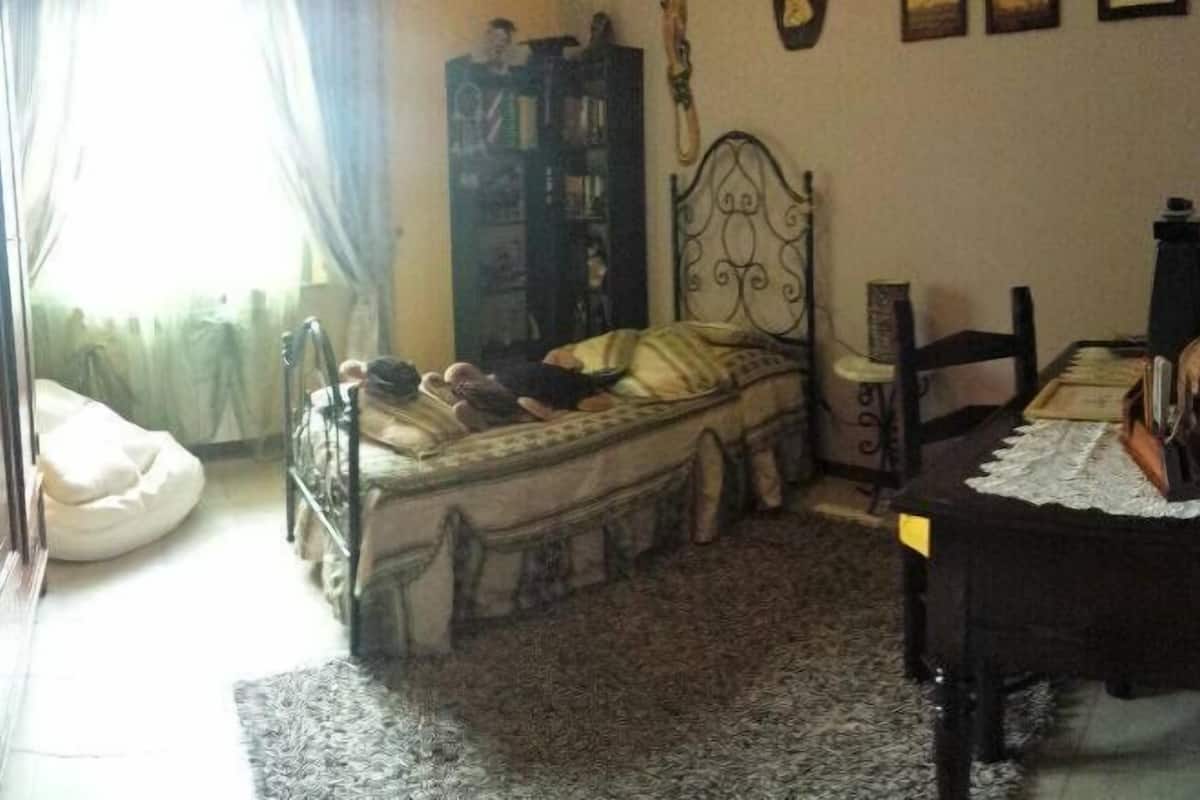 A bedroom with a bed, a wooden cabinet, a desk with a chair, and a window with curtains.