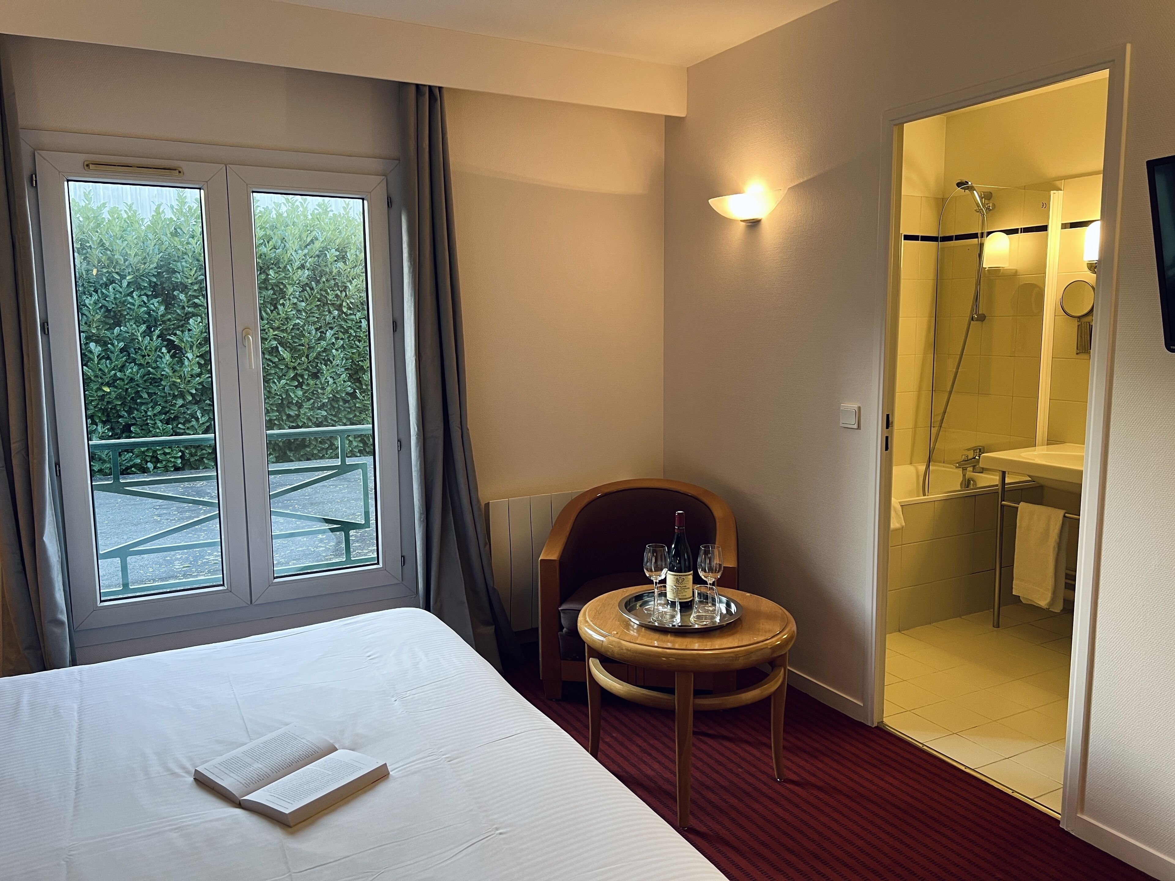 A hotel room with a bed, a small table with a wine bottle and glasses, a shower area, and a balcony with a view of greenery.