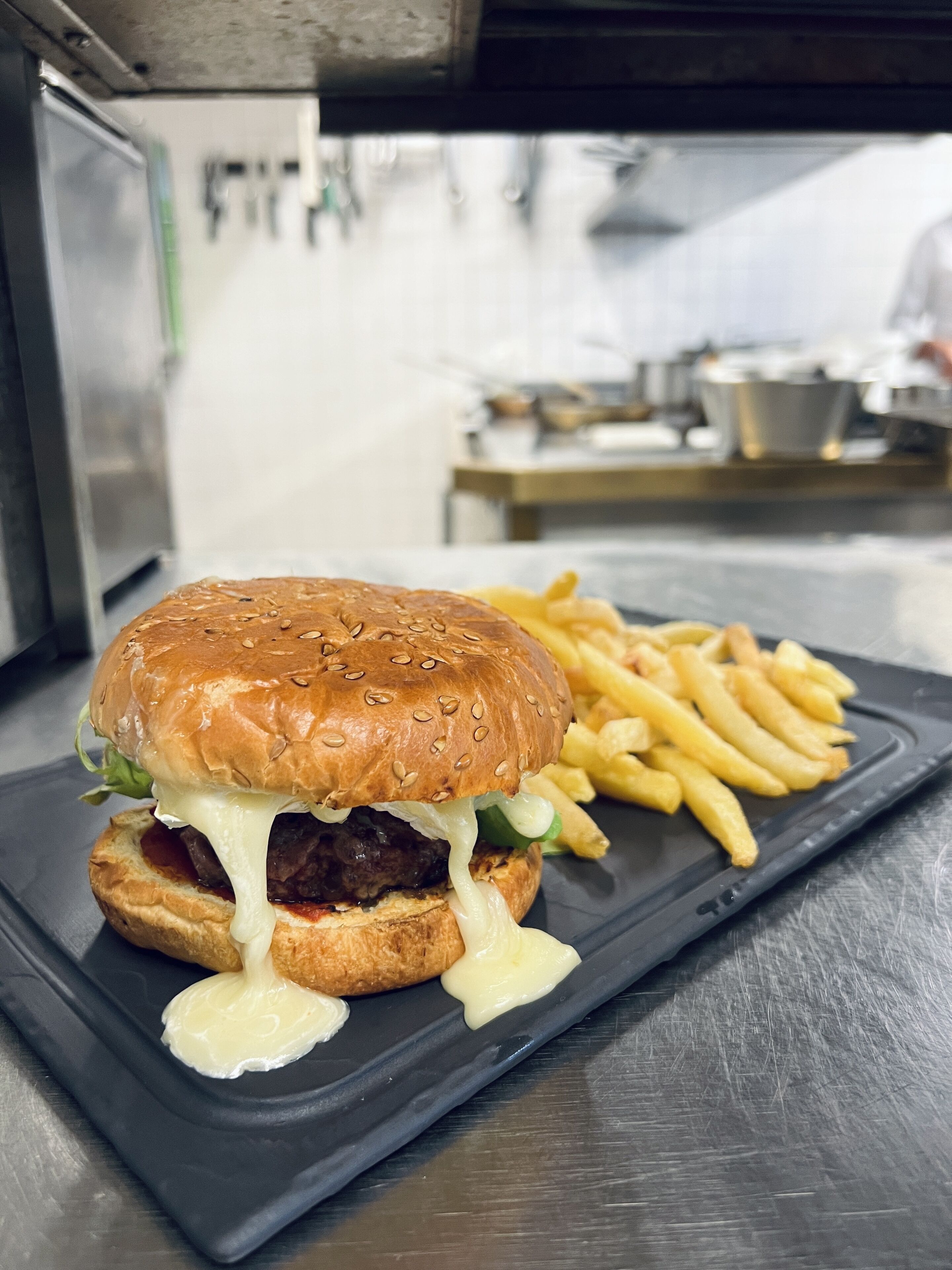 A burger with melted cheese and fries on a black plate in a kitchen setting.