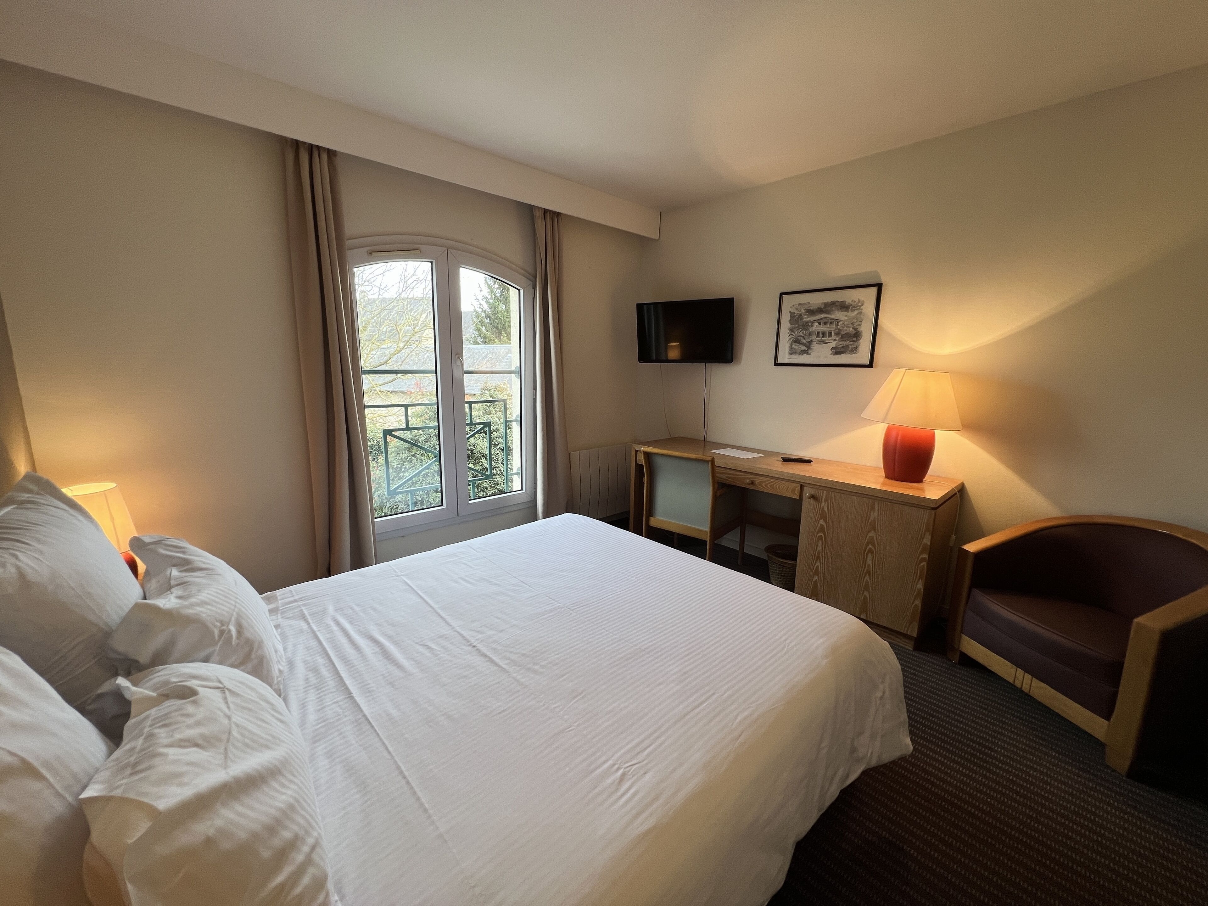 Ett hotellrum med en säng, ett skrivbord, en stol, en tv och ett fönster med gardiner.