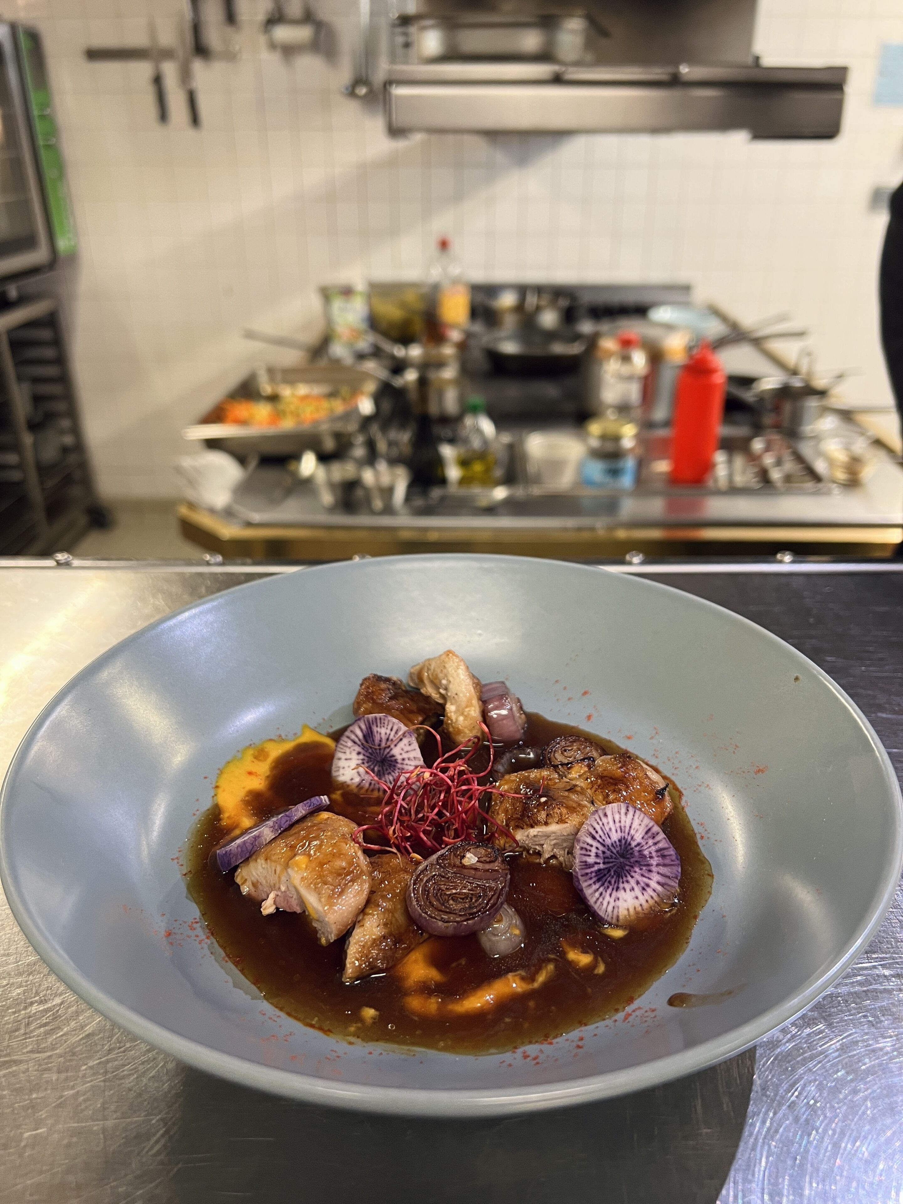 A bowl of food with a sauce, garnishes, and a kitchen in the background.