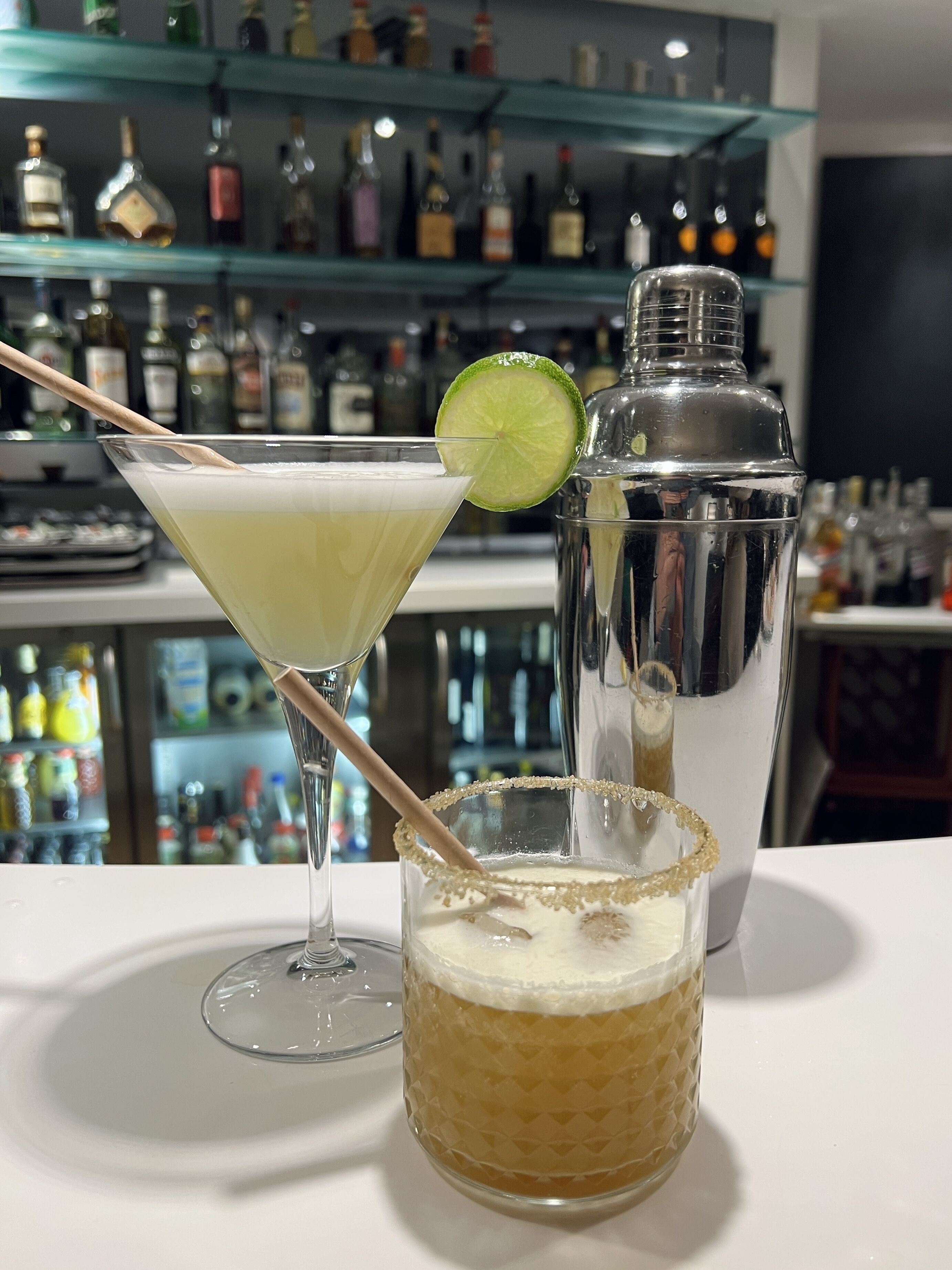 Un cocktail décoré d’une tranche de citron vert, posé sur un comptoir de bar, avec un shaker à côté.