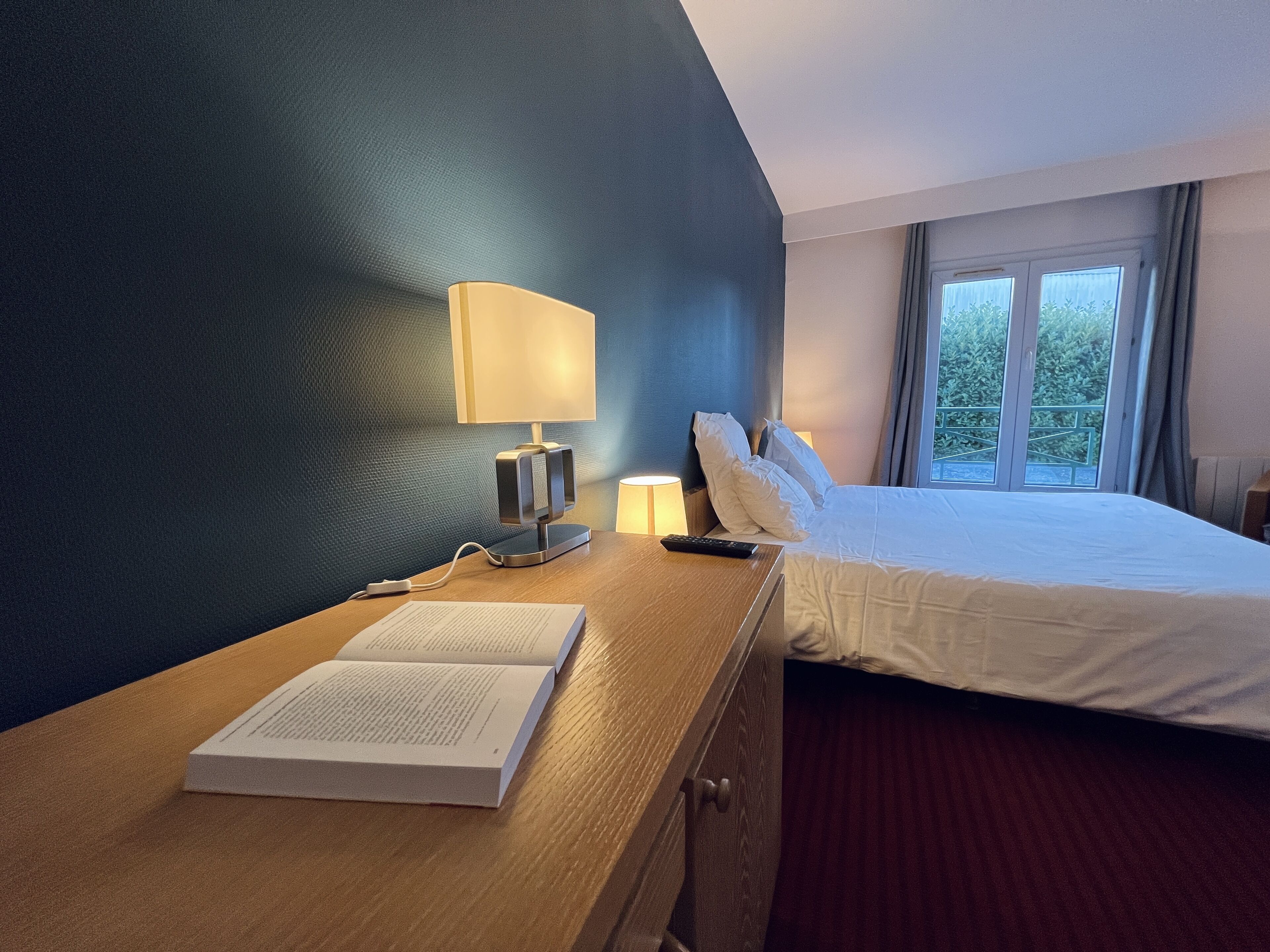 Et hotelværelse med en seng, et natbord med en lampe, en fjernbetjening og en åben bog.