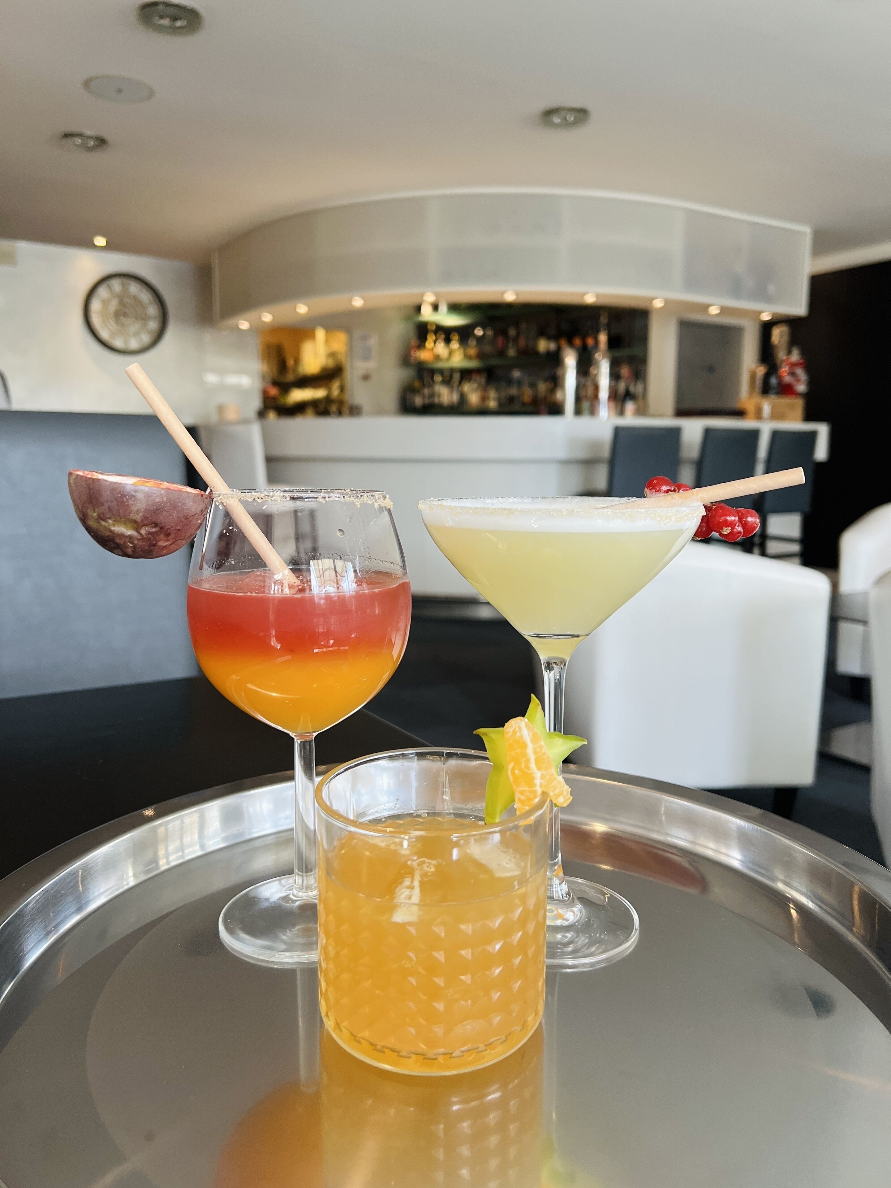 Three cocktails on a tray in a bar area with a modern interior and a bar stocked with bottles.