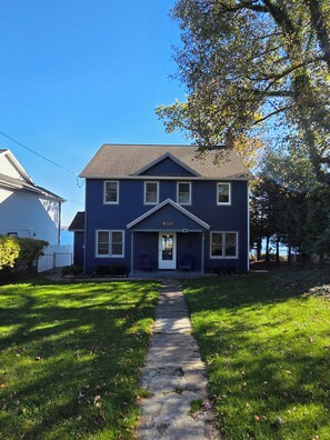 Une maison à deux étages avec un porche à l’avant, un chemin menant à l’entrée et un ciel bleu dégagé.
