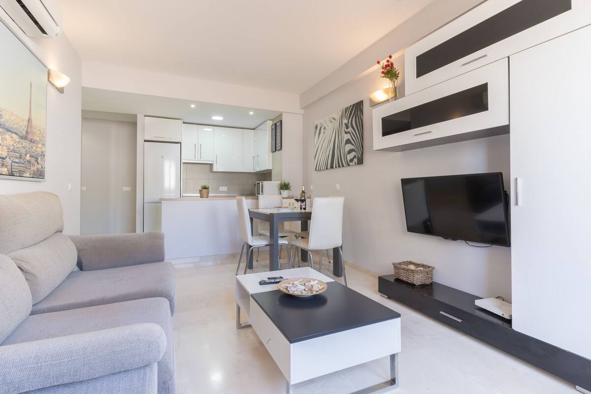 Un salon moderne avec un canapé, une table basse et un téléviseur. On aperçoit à l’arrière-plan un coin repas avec des chaises et une table.