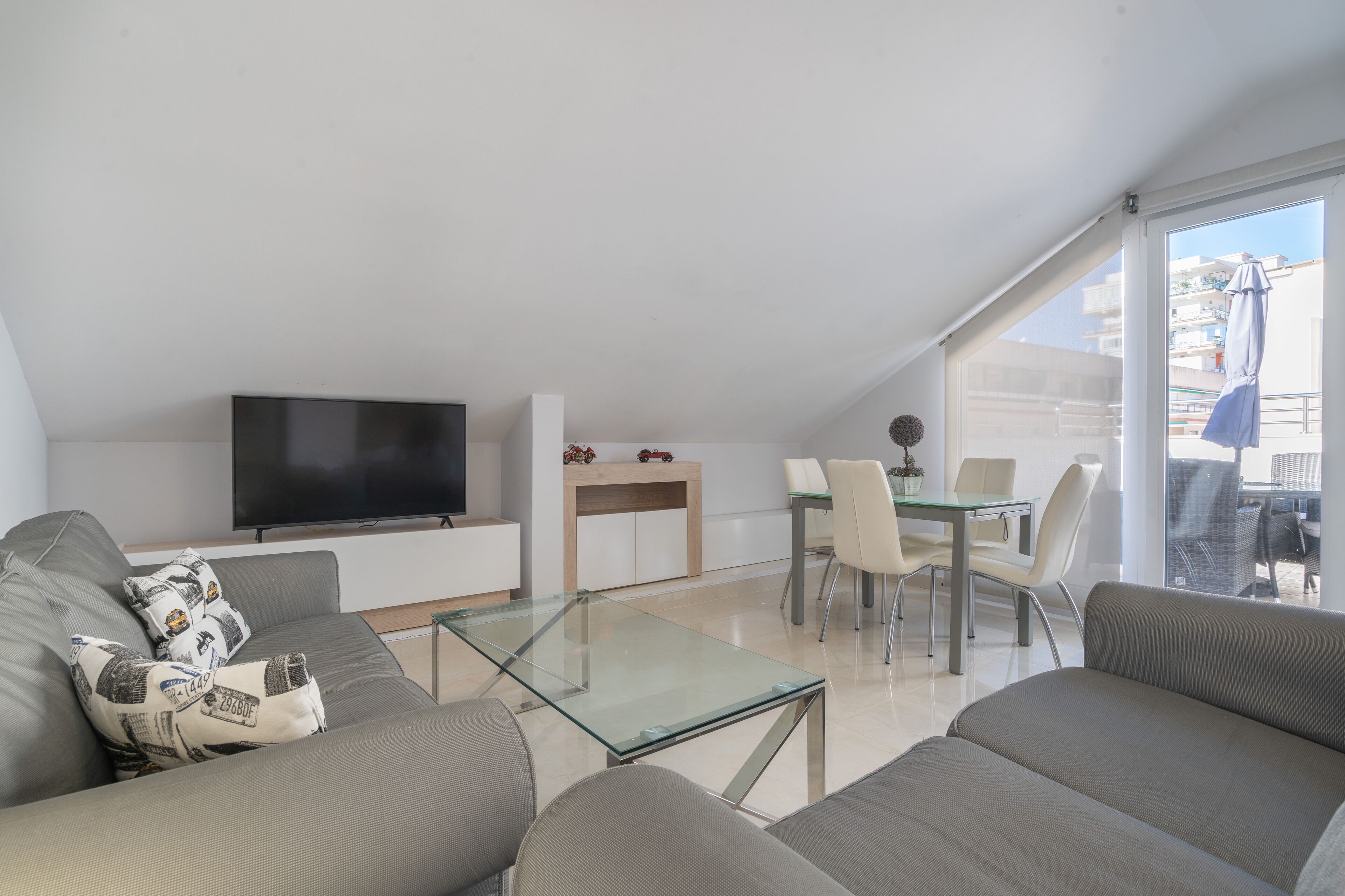 Un salon moderne avec une table basse en verre, des canapés gris, un téléviseur à écran plat, un coin repas avec une table et des chaises en verre, et un balcon offrant une vue sur les immeubles.