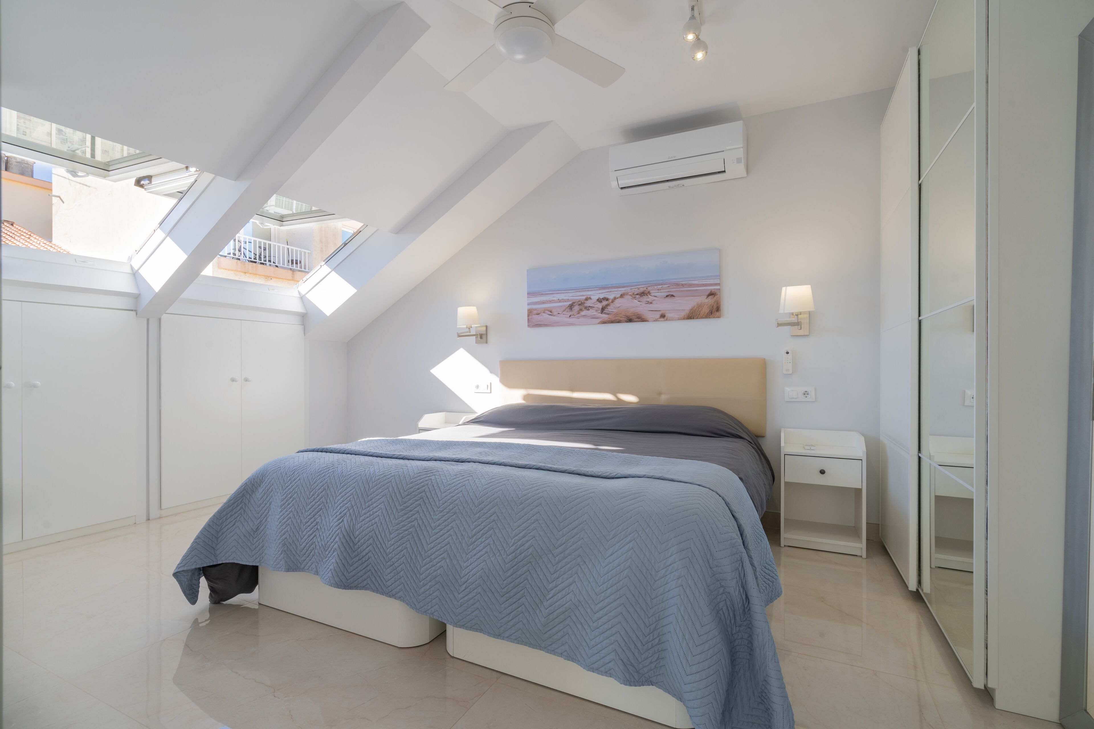 Un dormitorio moderno con tragaluz, una cama con colcha azul, una mesita de noche y una lámpara de pared.