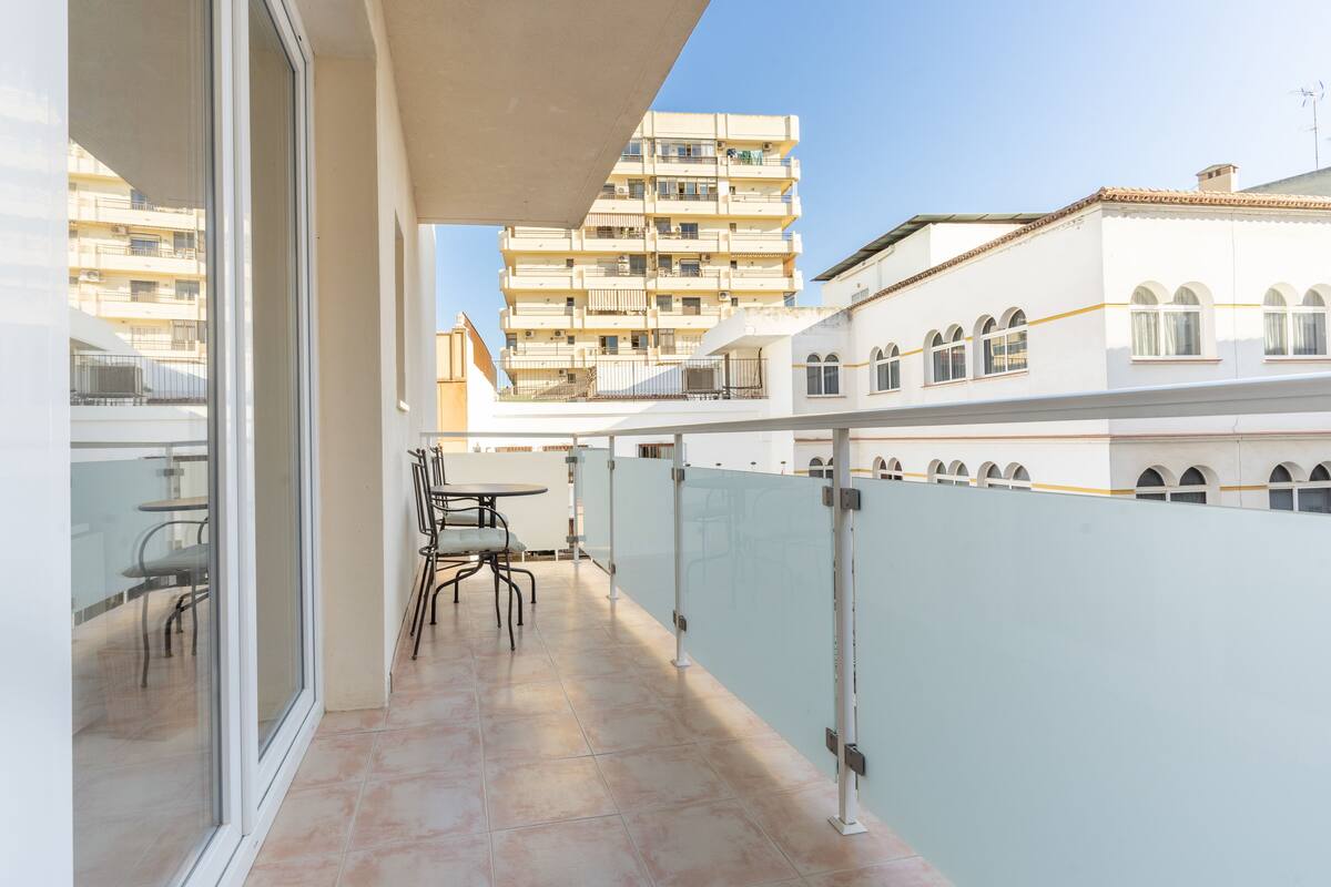 Un balcon avec une porte vitrée, une table et des chaises. On aperçoit plusieurs étages d’immeubles en arrière-plan.