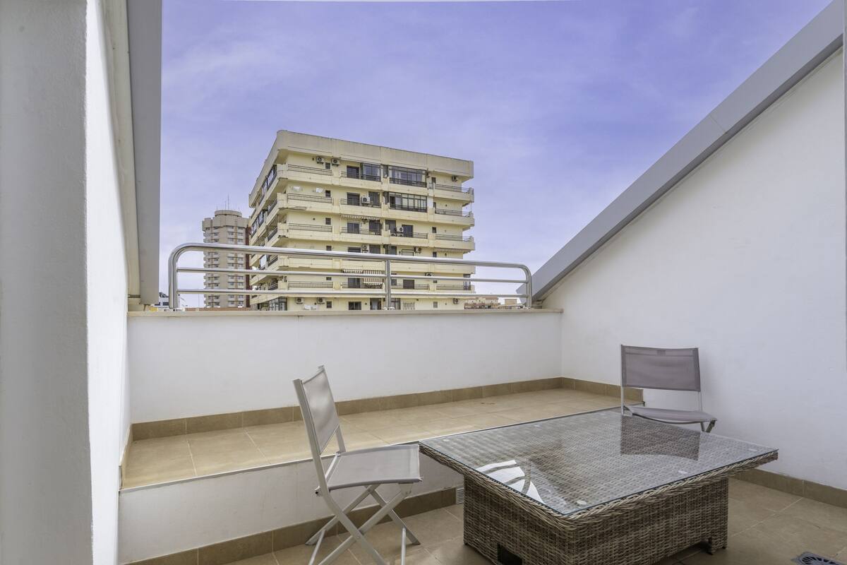 Un balcon avec une table et des chaises en verre, une vue sur un immeuble à plusieurs étages et un mur blanc.