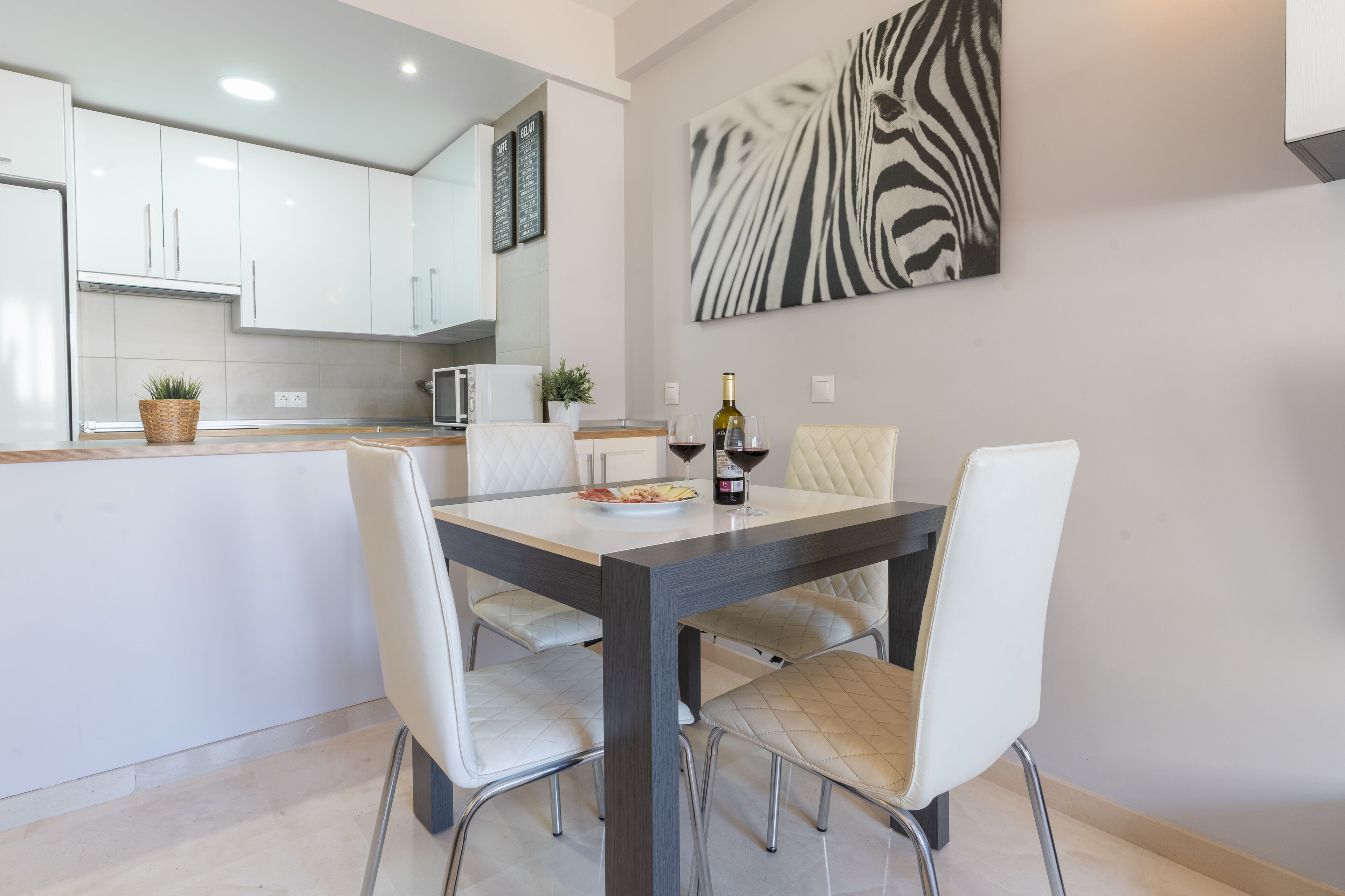 A modern kitchen with white cabinets, a dining table with four chairs, and a zebra print artwork on the wall.