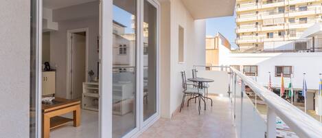 A balcony with a dining table and chairs, a sliding glass door leading to a kitchen, and a view of a multi-story building.