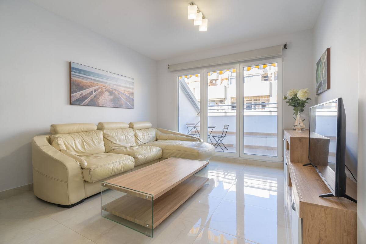 Un salon moderne avec un canapé en cuir beige, une table basse en verre, un téléviseur à écran plat et une porte-fenêtre coulissante donnant sur un balcon extérieur.
