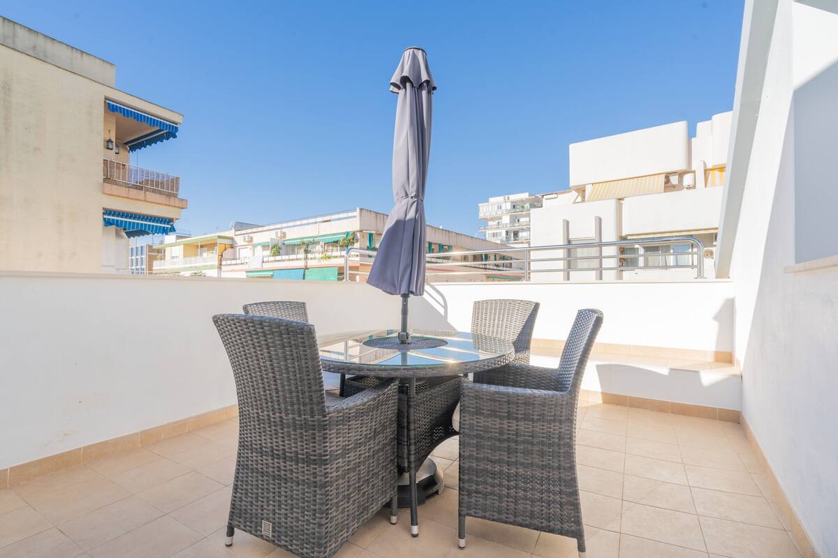 Une table ronde entourée de quatre chaises en osier, et un parasol gris, le tout sur un balcon d’où l’on a une vue dégagée sur les bâtiments et une piscine en arrière-plan.