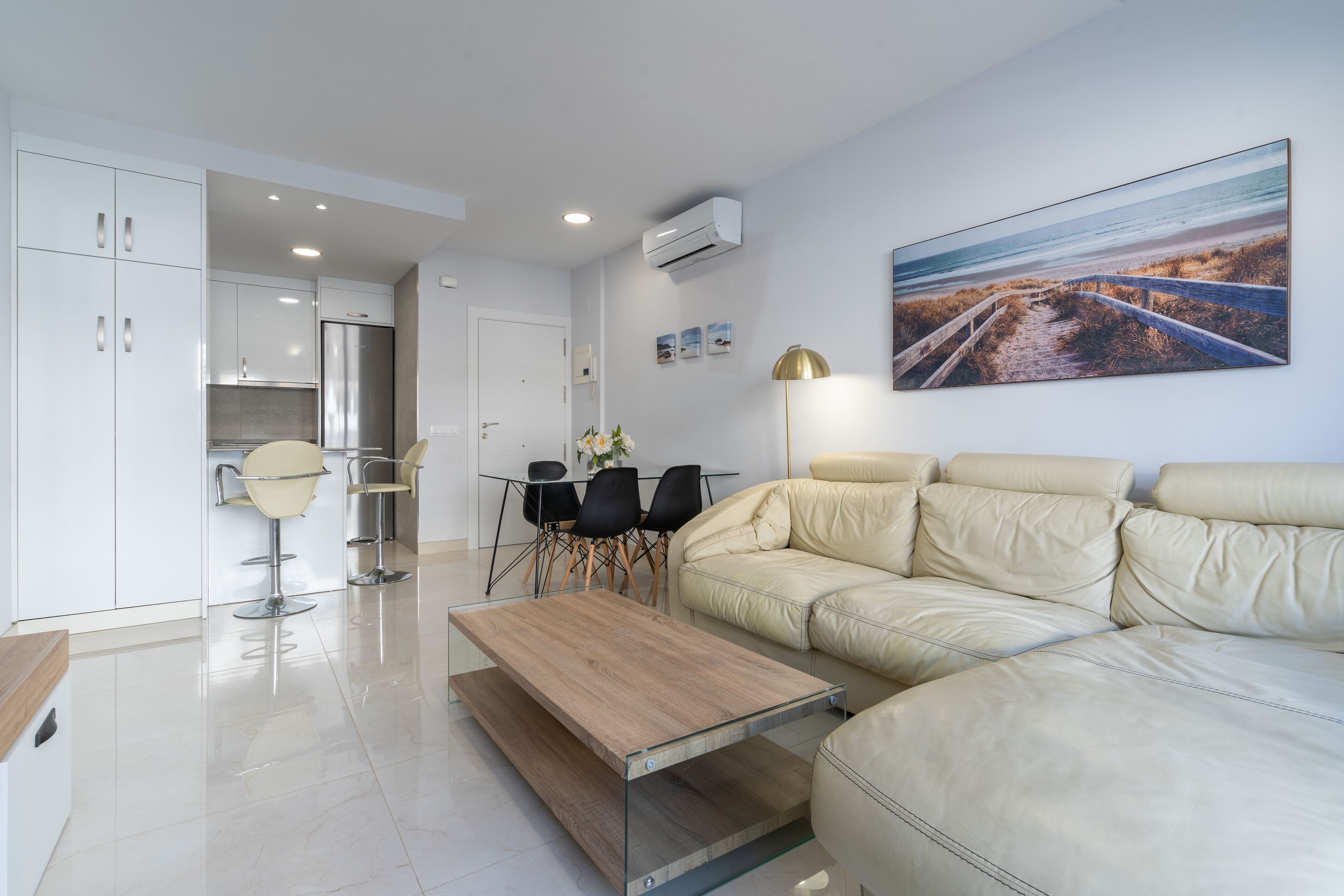A modern living room with a beige sofa, a glass coffee table, and a dining area with a white cabinet and black chairs.