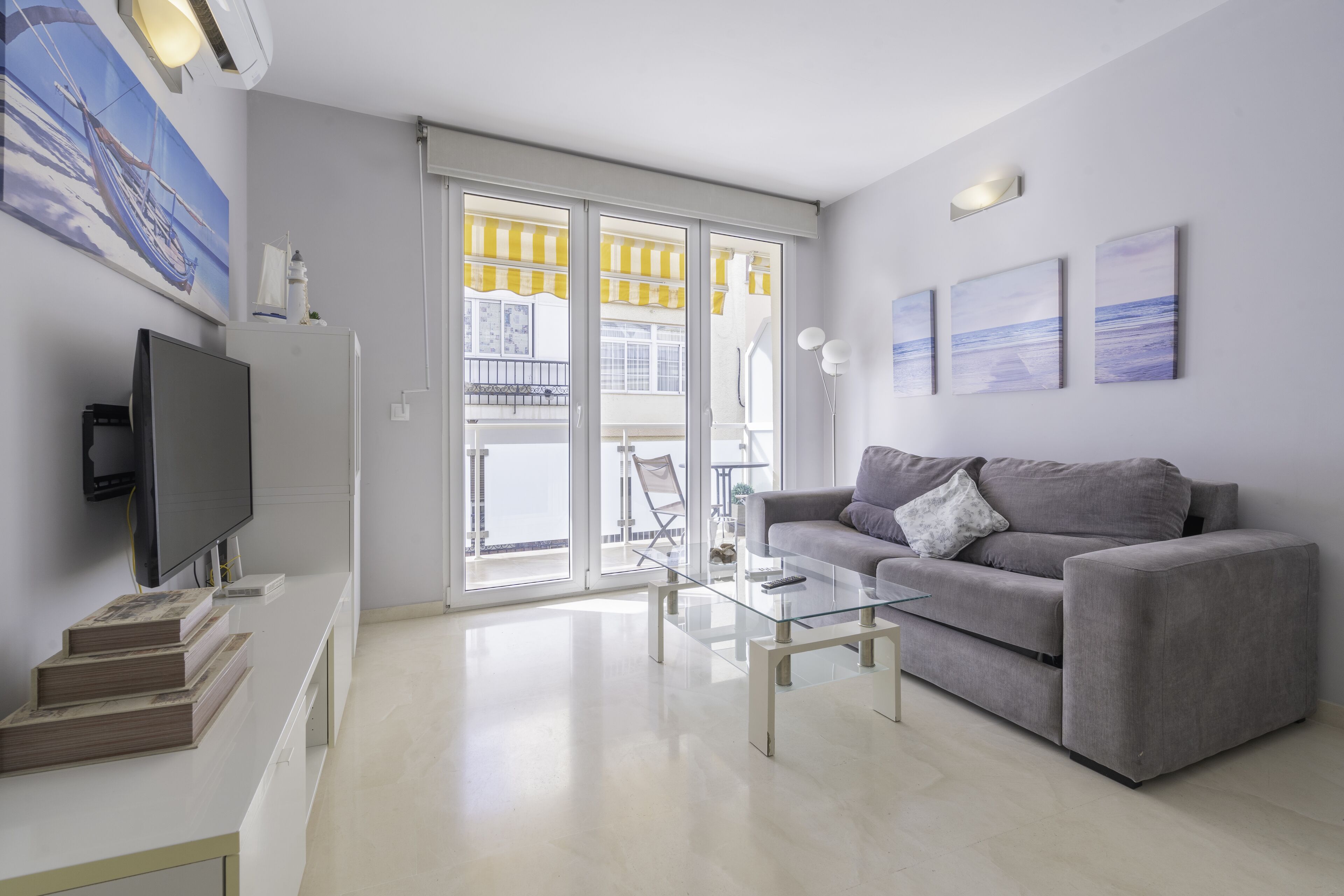 A modern living room with a grey sofa, a glass coffee table, and a flat-screen TV. There is a balcony with yellow awnings and a view of the sea.