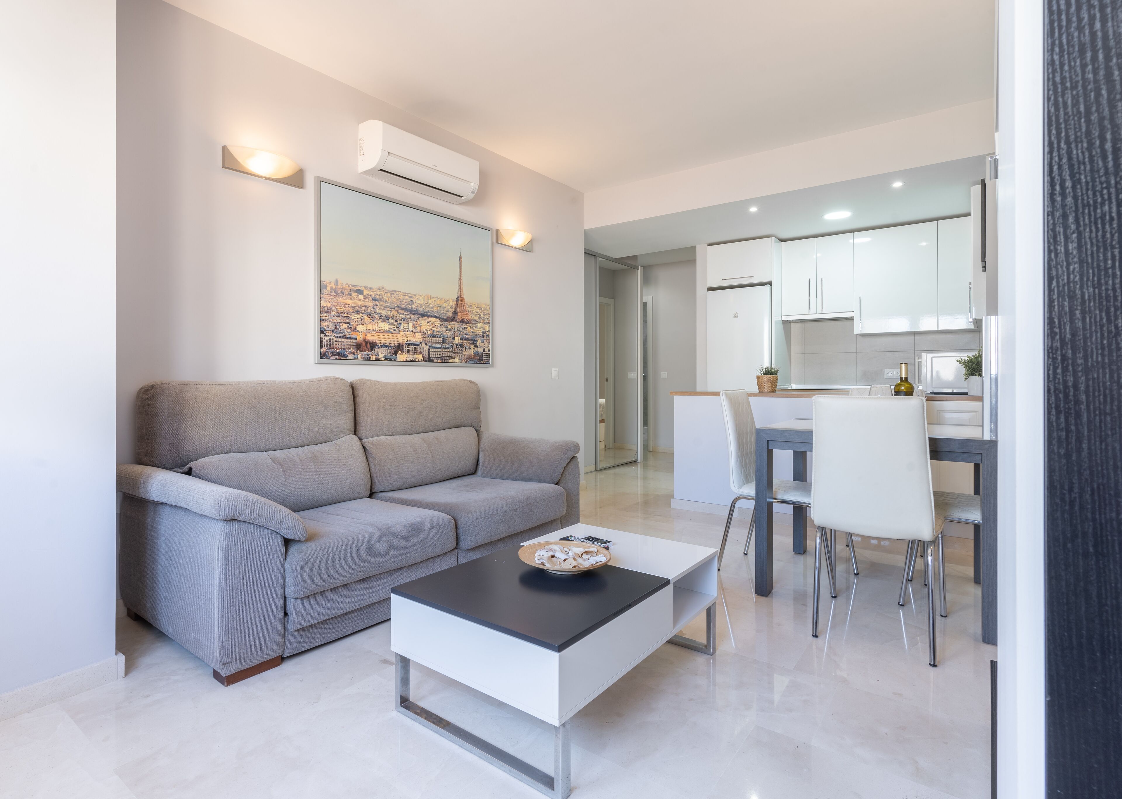 A modern living room with a grey sofa, a coffee table, and a dining area with a white table and chairs.