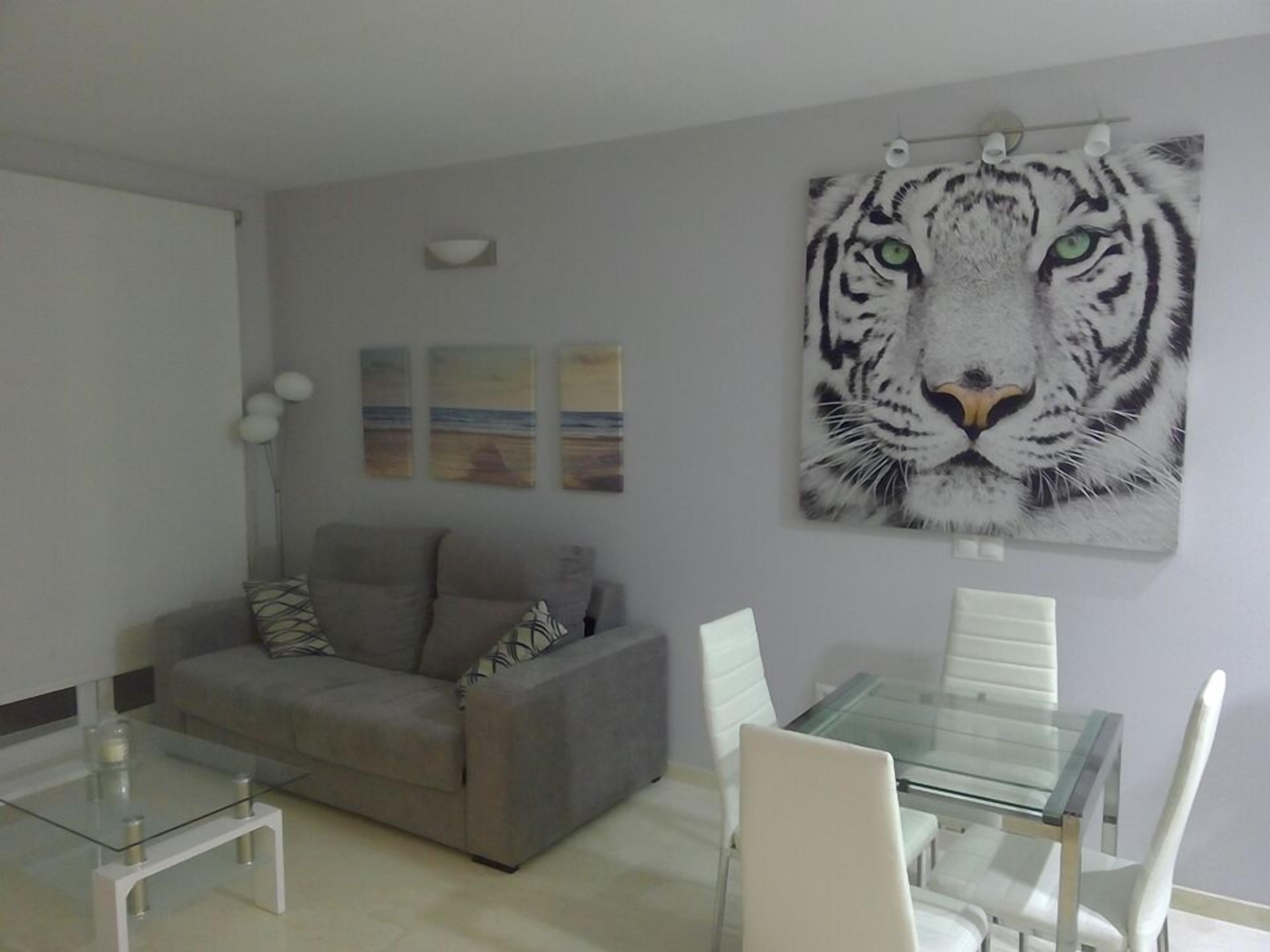 A living room with a grey sofa, a glass coffee table, and a glass dining table with white chairs. There is a large tiger painting on the wall.
