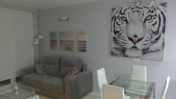 A living room with a grey sofa, a glass coffee table, and a glass dining table with white chairs. There is a large tiger painting on the wall.