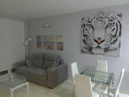 A living room with a grey sofa, a glass coffee table, and a glass dining table with white chairs. There is a large tiger painting on the wall.